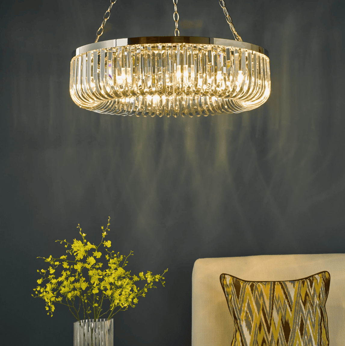 Dar lighting chandelier hanging above a vase with yellow flowers and a beige chair with a patterned pillow against a dark background.