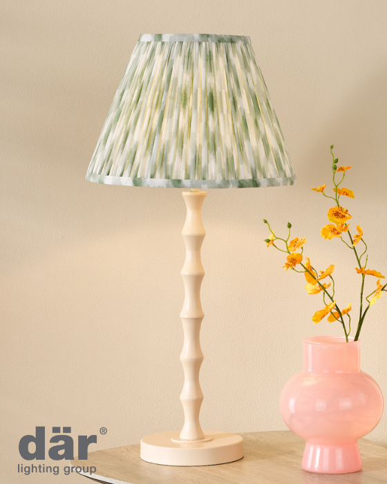 Table lamp with pleated green shade and cream wooden base next to a pink vase with yellow flowers on a beige background.
