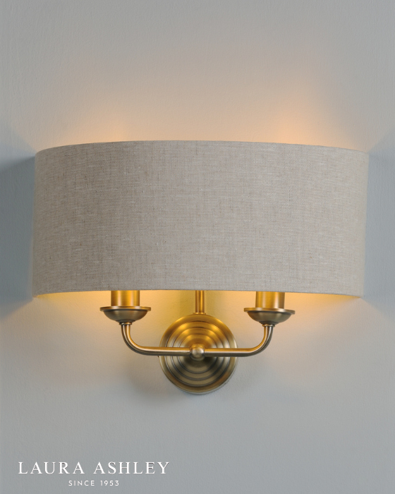 Antique brass wall light with two arms and natural shade on a pale grey background with Laura Ashley logo, bottom left. 