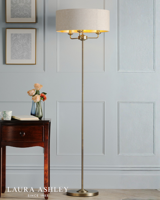 Floor lamp in antique brass finish with natural shade and 'Laura Ashley' on a pale blue panelled background with logo in white, bottom left. 