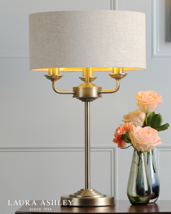 Classic antique brass table lamp with natural shade on pale grey background with Laura Ashley logo, bottom left.